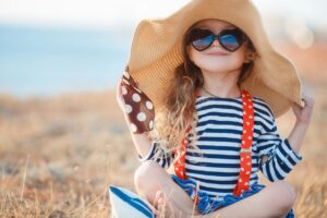 A little girl sitting on the ground wearing sunglasses and a hat.